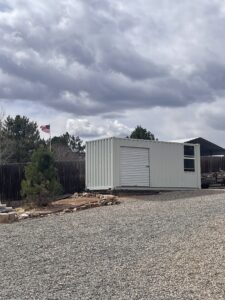 Shipping container being used for storage in a residential backyard with an added roll up door and window.  White clouds, evergreen trees and the American flag stand in the background.