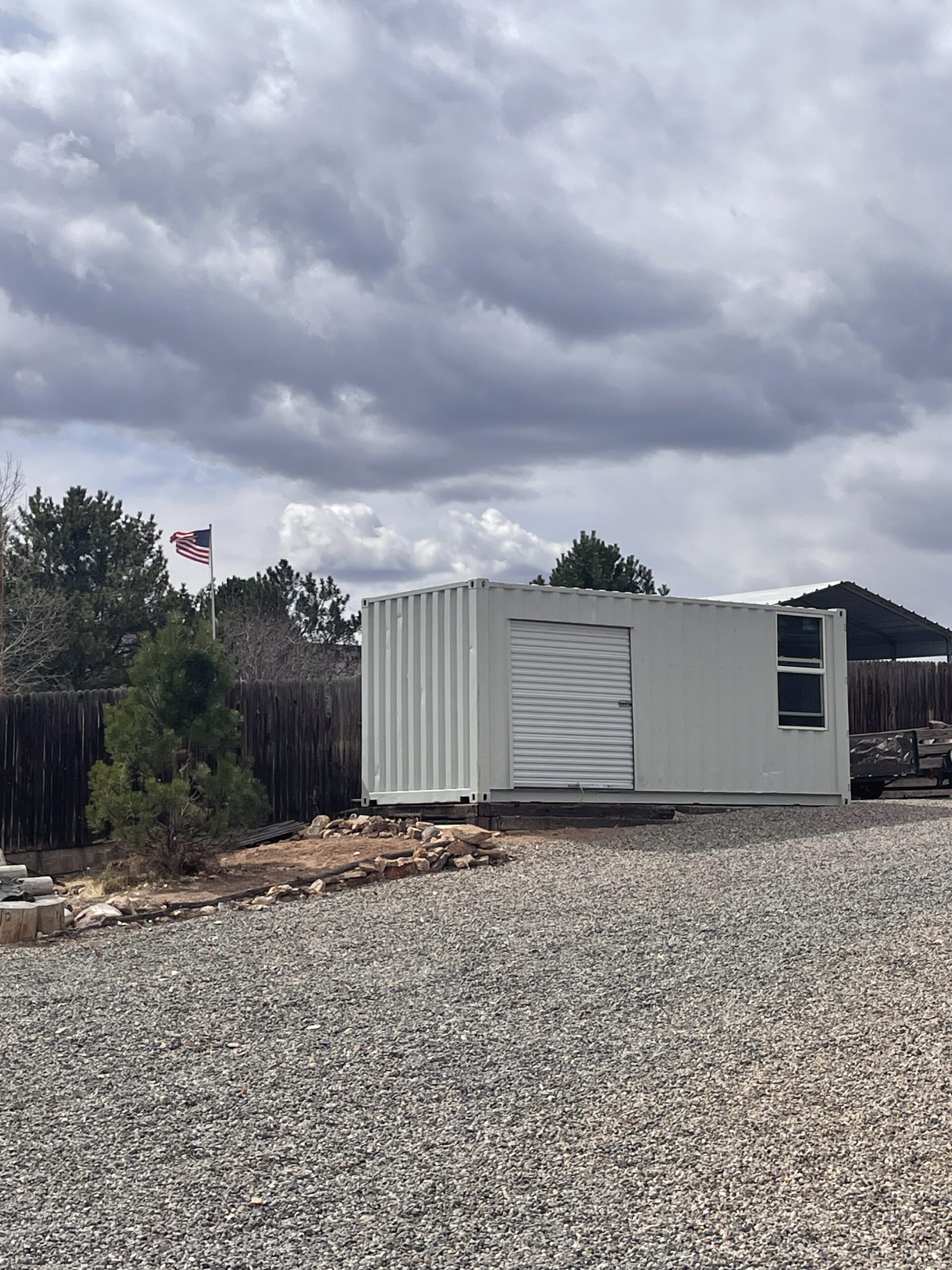 Shipping container being used for storage in a residential backyard with an added roll up door and window. White clouds, evergreen trees and the American flag stand in the background.