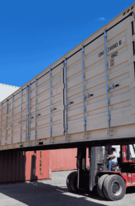 A tan, open side storage container is being lifted by a forklift approximately 5 ft off the ground.  The long side of the storage container with closed open side doors face the camera.